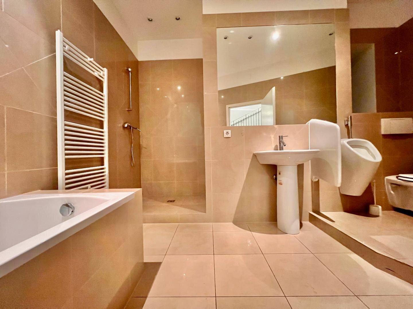 Modernes Badezimmer mit beigefarbenen Fliesen, Badewanne, Duschbereich und wandhängendem WC.