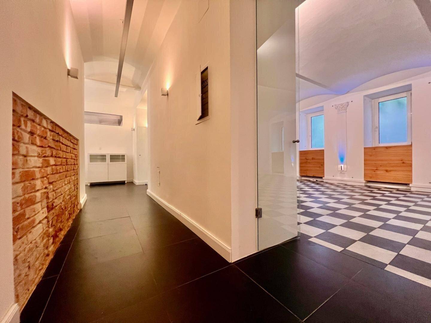 Heller Flur mit freiliegender Ziegelwand, dunklem Fliesenboden und Glasabtrennung zu einem Raum mit Schachbrettmuster.