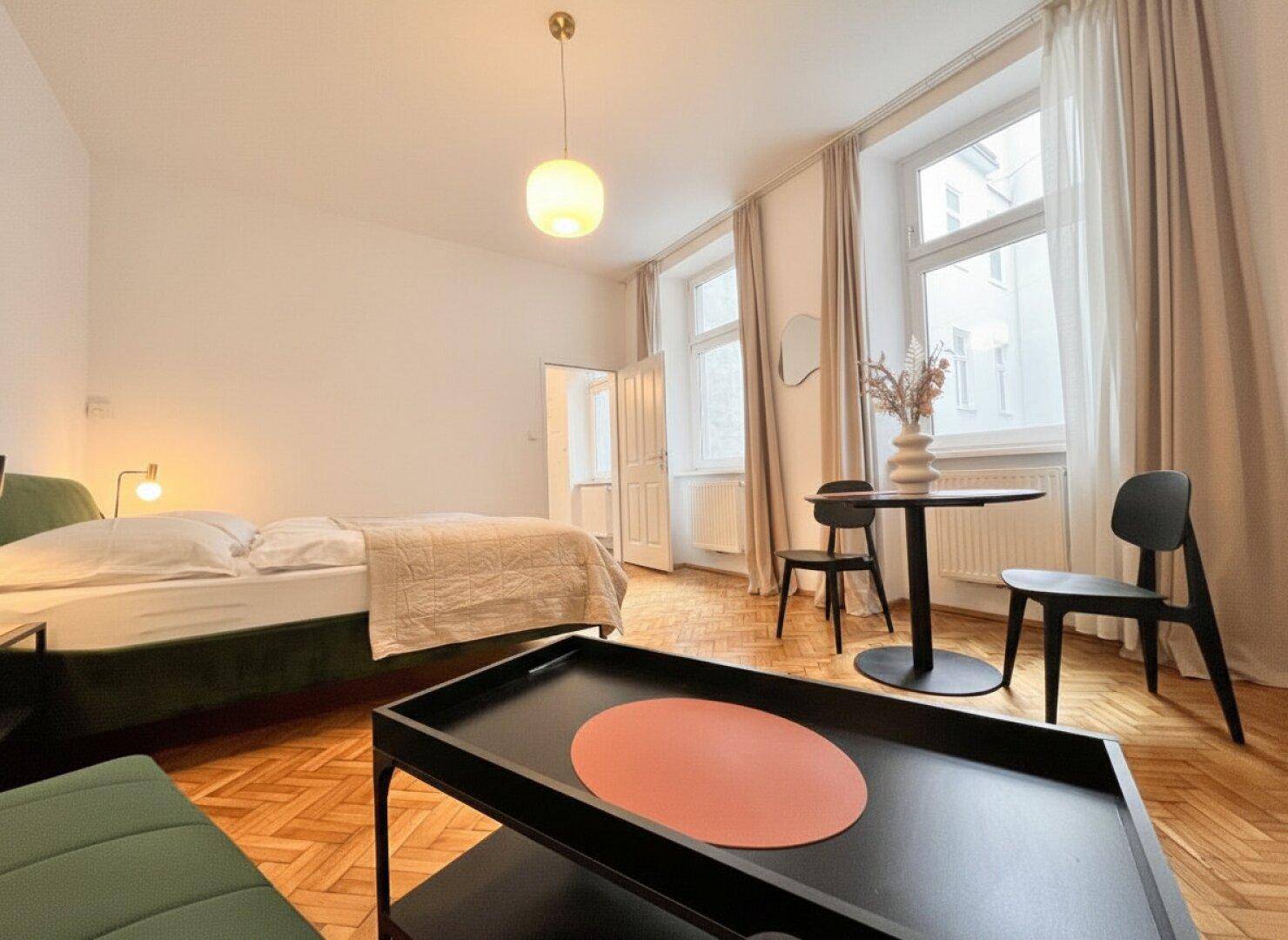 Gemütlicher Wohnbereich mit Parkettboden, großen Fenstern und einem kleinen Esstisch.