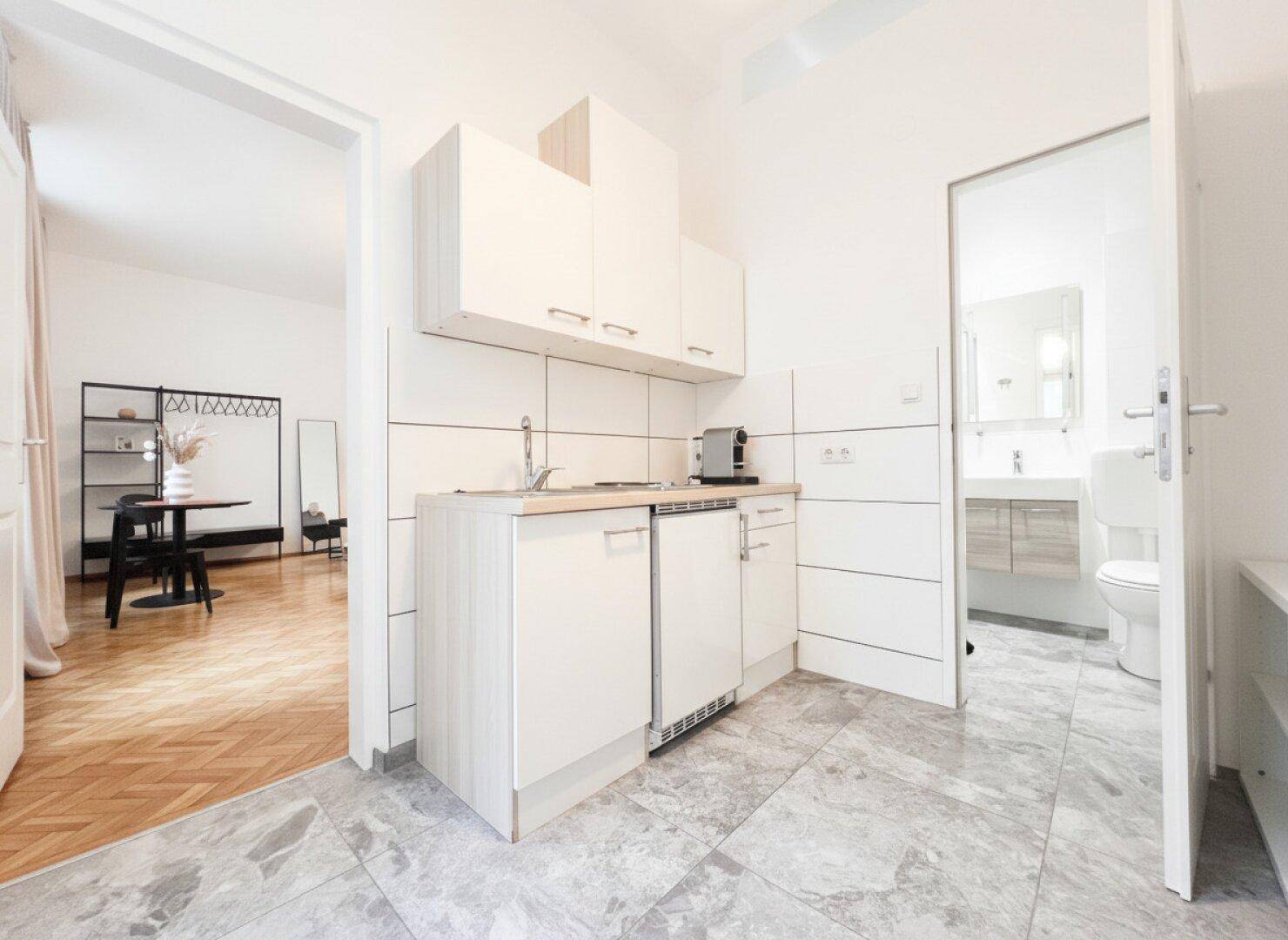 Kompakte Küchenzeile mit weißen Schränken und Zugang zum Wohnbereich und Bad.