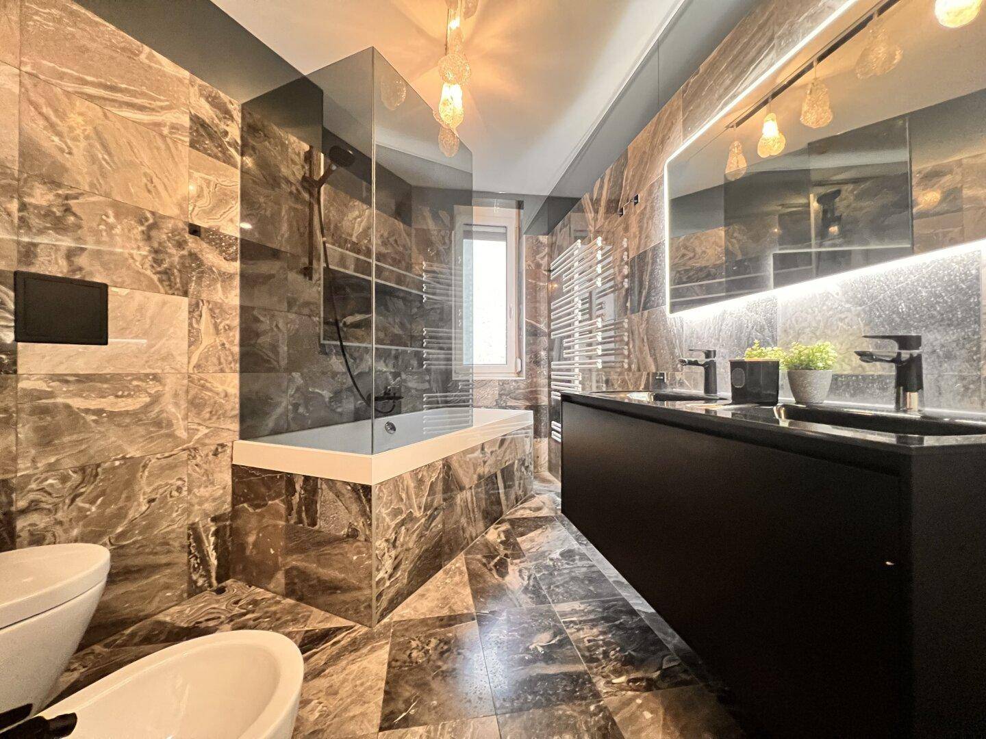 Elegantes Badezimmer mit dunklen Marmorfliesen, Doppelwaschbecken und beleuchtetem Spiegel.