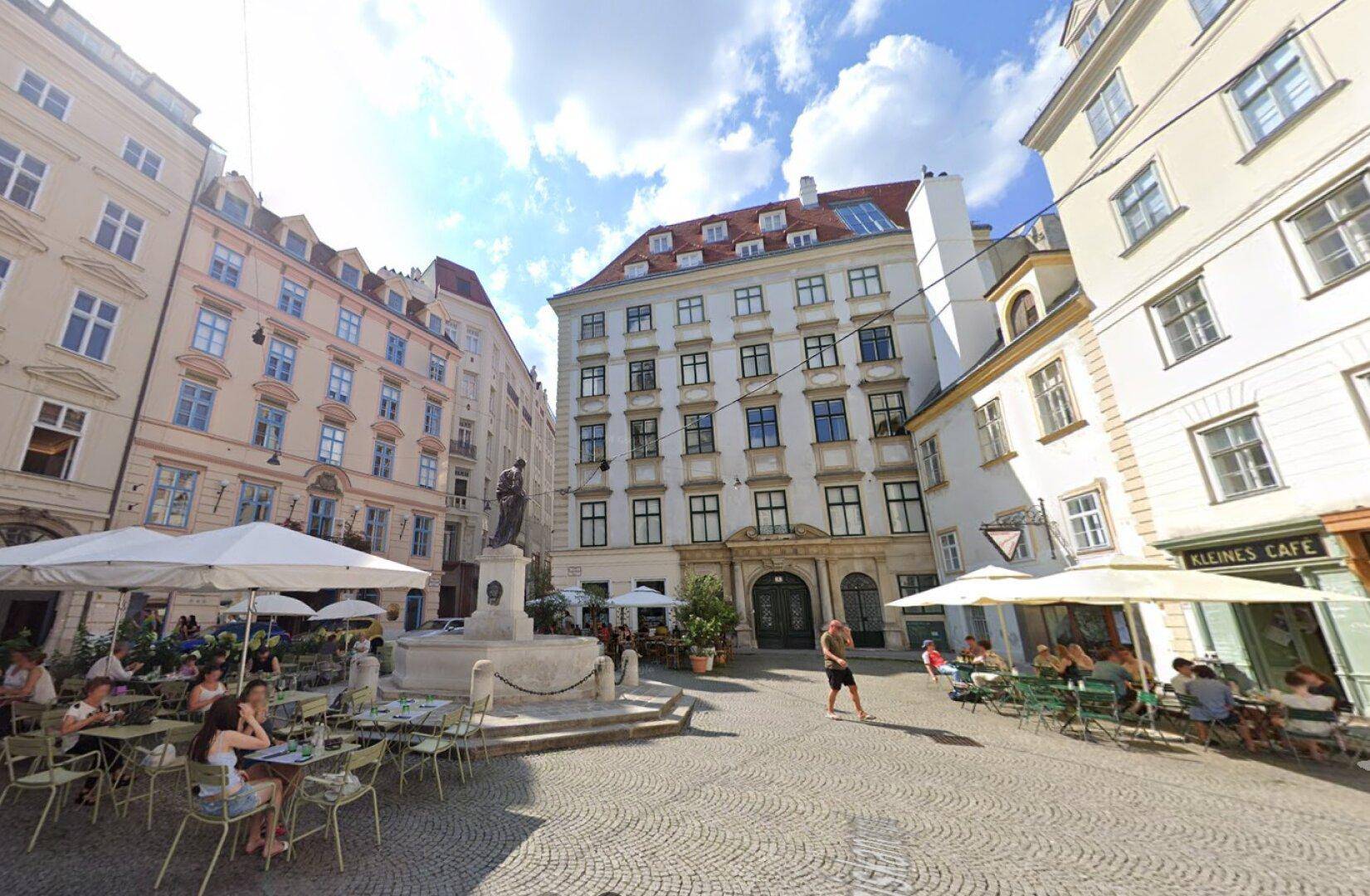 Malerischer Stadtplatz mit Cafés, Brunnen und historischen Gebäuden unter blauem Himmel.