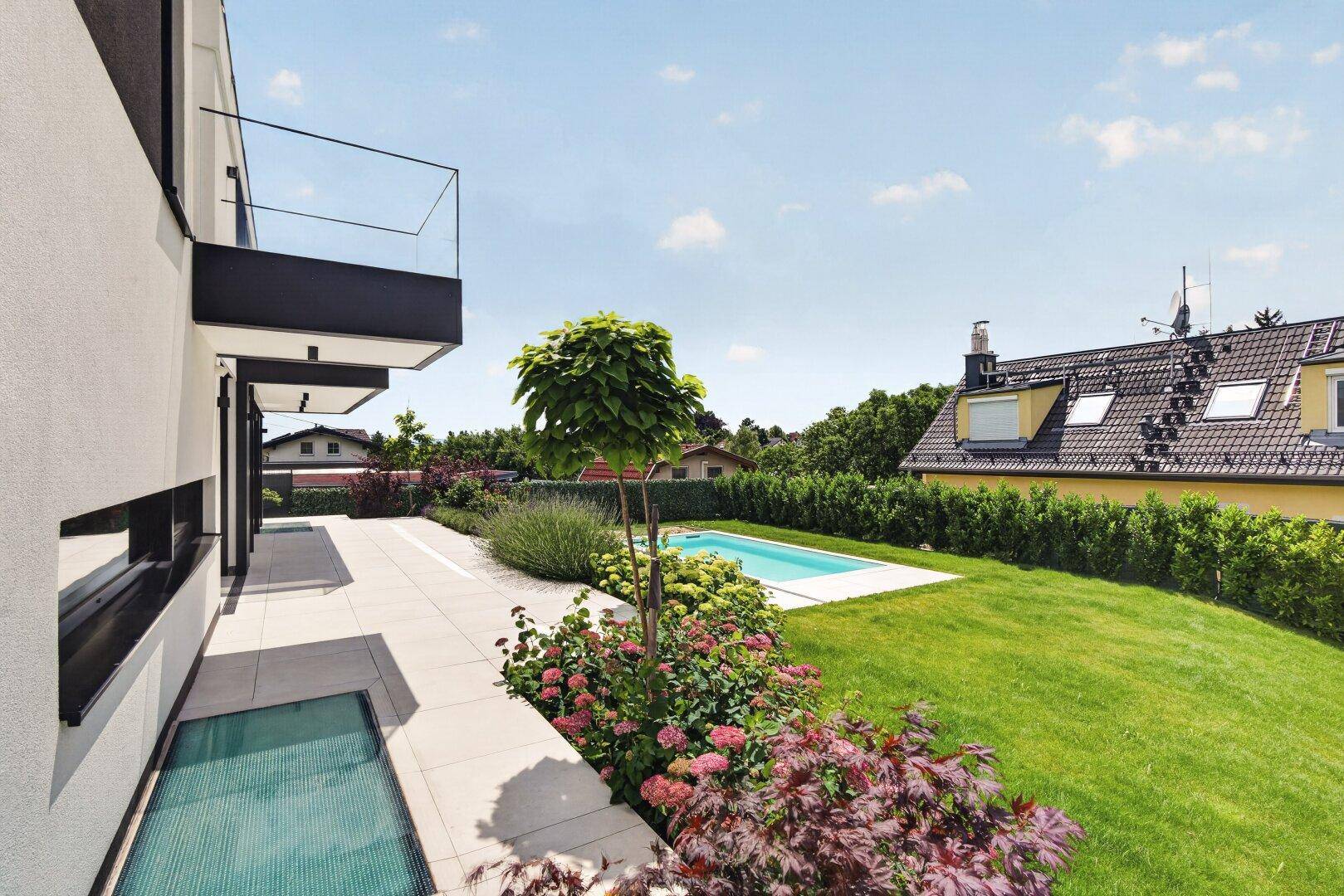 Seitlicher Blick auf das Haus mit gepflegtem Rasen, Blumenbeeten und einem kleinen Pool im Hintergrund.