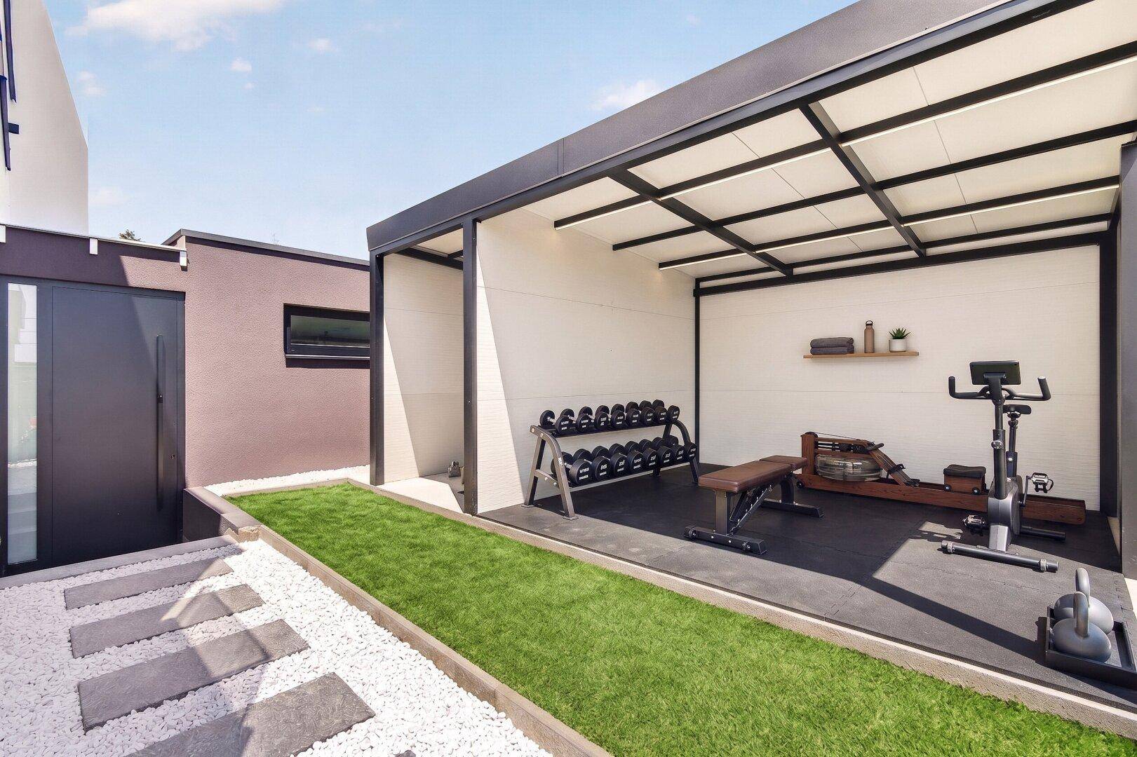 Überdachter Außenbereich, eingerichtet als Fitnessstudio mit verschiedenen Trainingsgeräten und Hanteln.