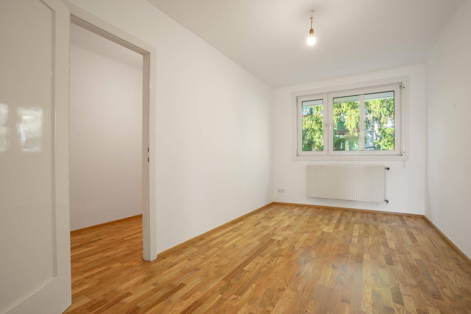 Helles Schlafzimmer mit Parkettboden und Fenster zum grünen Außenbereich, in gutem Zustand.