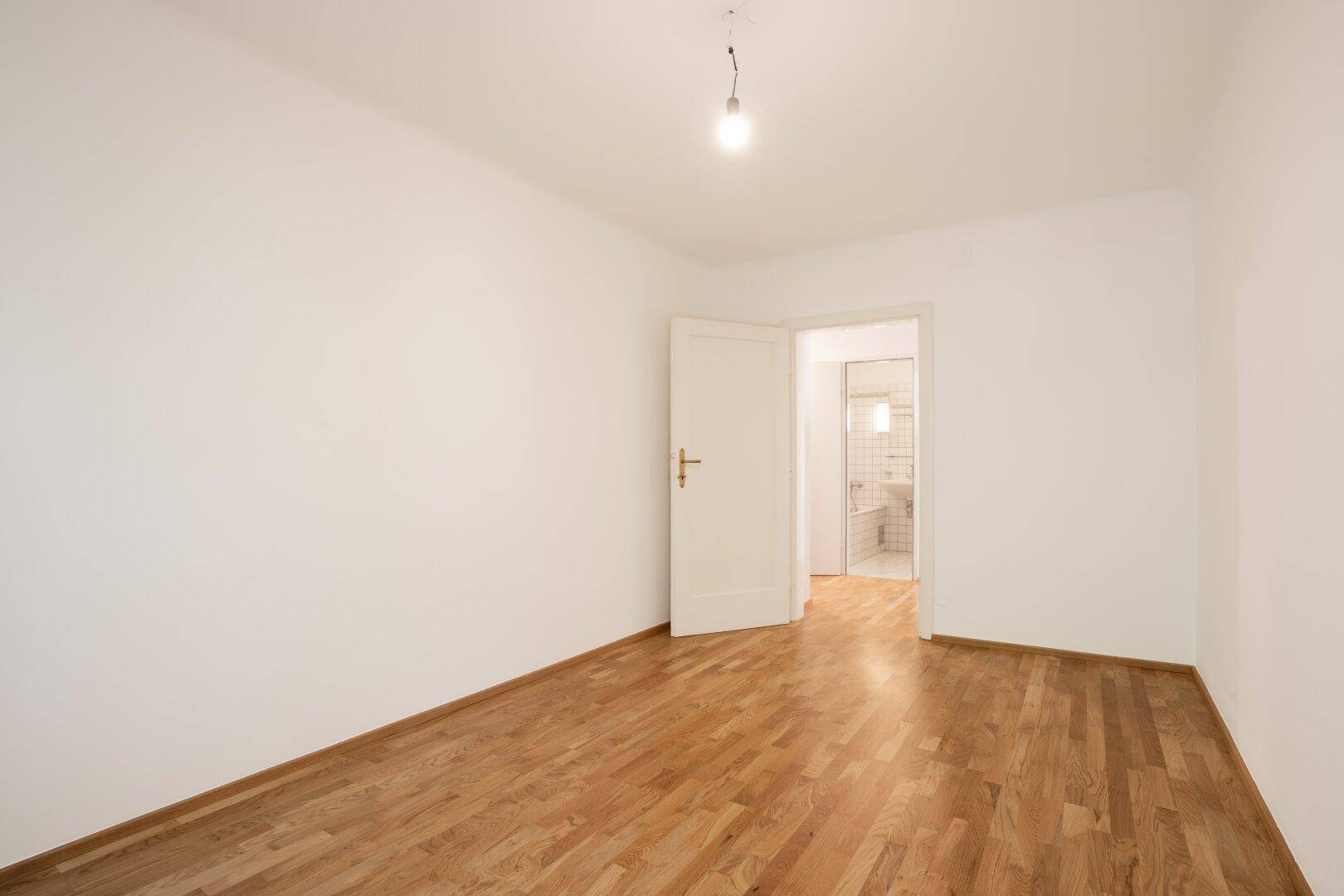 Geräumiges Zimmer mit Parkettboden und Zugang zu einem angrenzenden Badezimmer, hell und sauber.