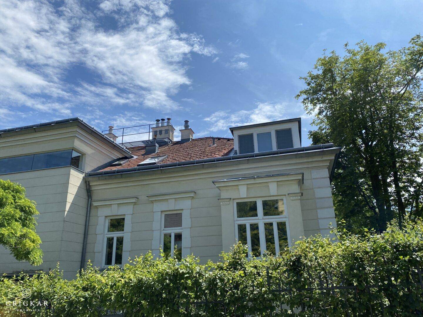 Klassische Gebäudefassade mit rotem Ziegeldach, Dachgauben und grüner Hecke im Vordergrund.