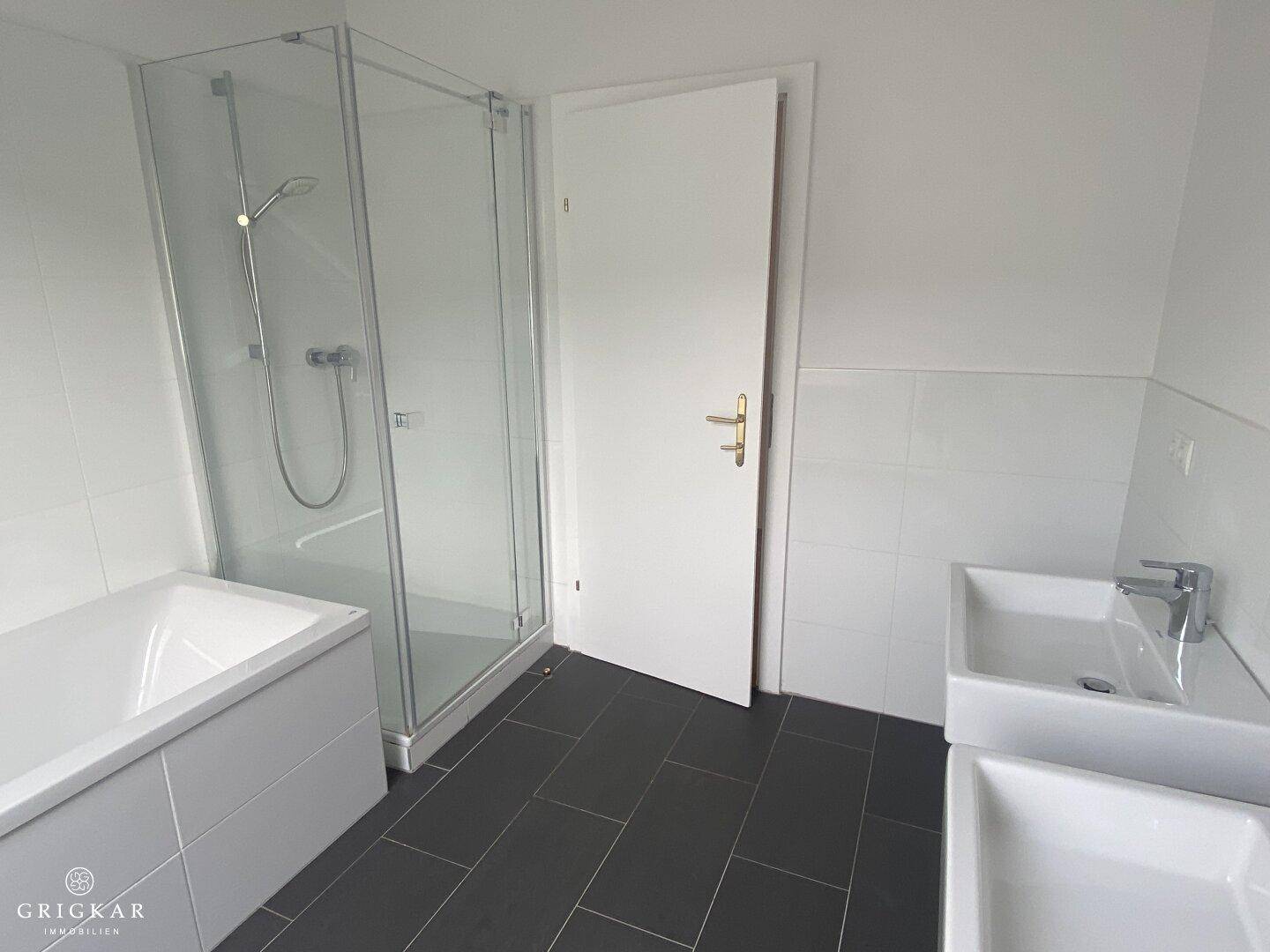Badezimmer mit moderner Duschkabine, Badewanne und Waschbecken auf dunklem Fliesenboden.