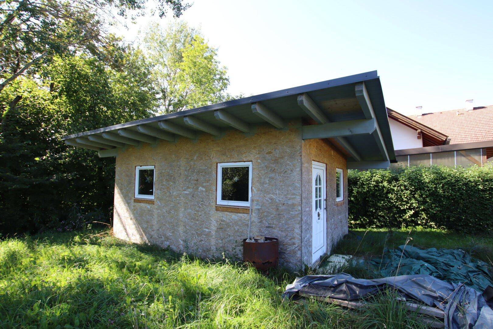 Kleiner Holzschuppen mit Pultdach und weißen Fenstern, umgeben von Gras und Bäumen.