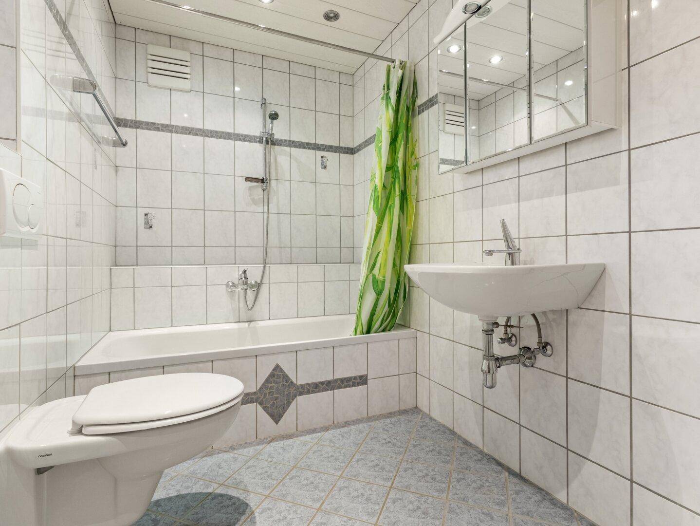 Helles Badezimmer mit weißen Fliesen, Badewanne mit Duschfunktion und Waschbecken.
