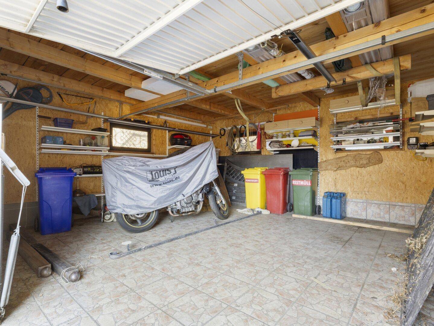 Garage mit Holzbalken, Fliesenboden und Stauraum für Fahrzeuge und Mülltonnen.