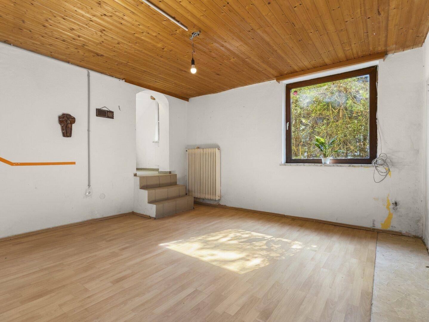 Wohnzimmer mit Holzbalkendecke, Laminatboden und einem Fenster mit Blick ins Grüne.
