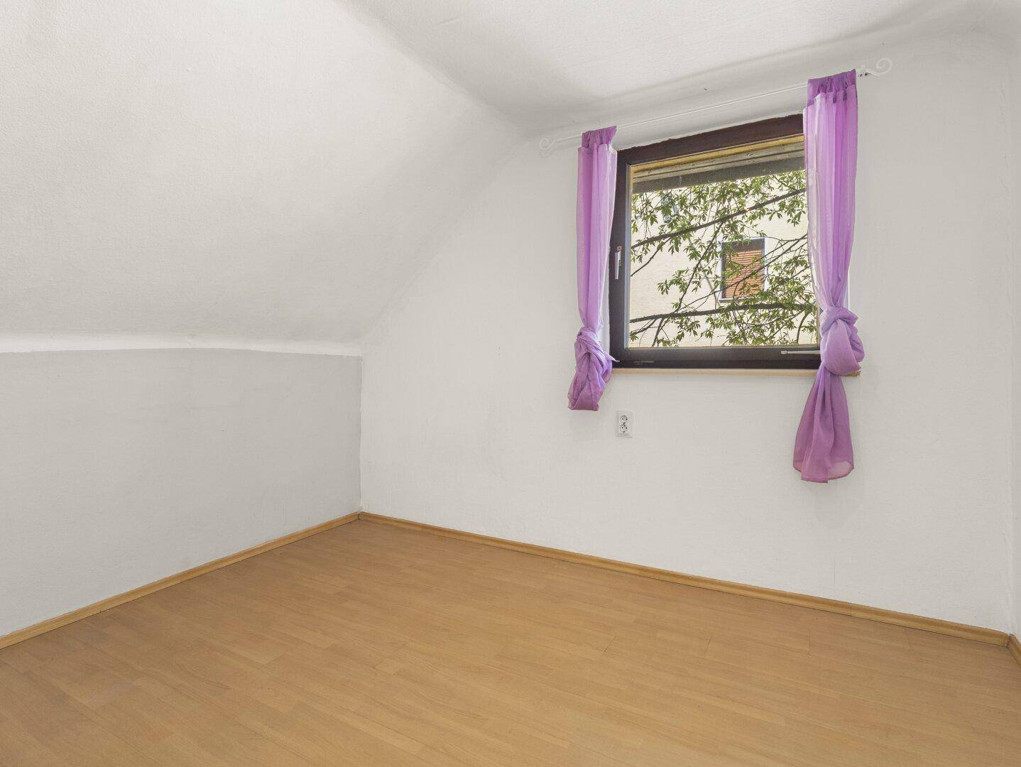 Schlafzimmer im Dachgeschoss mit Laminatboden, schrägen Wänden und einem Fenster mit lila Vorhängen.