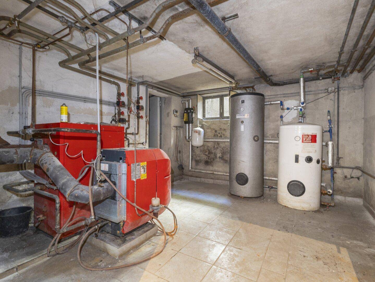 Heizungsraum im Keller mit Heizkessel, Warmwasserspeichern und sichtbaren Rohrleitungen.