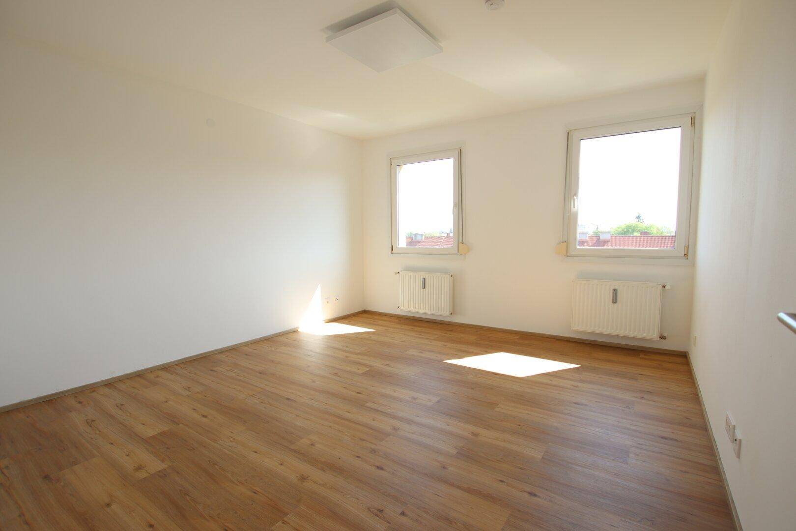 Helles Zimmer mit Laminatboden, weißen Wänden und zwei Fenstern mit Blick nach draußen.