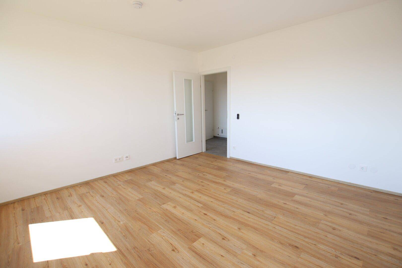 Lichtdurchflutetes Zimmer mit Laminatboden und weißer Tür, die zu einem anderen Raum führt.