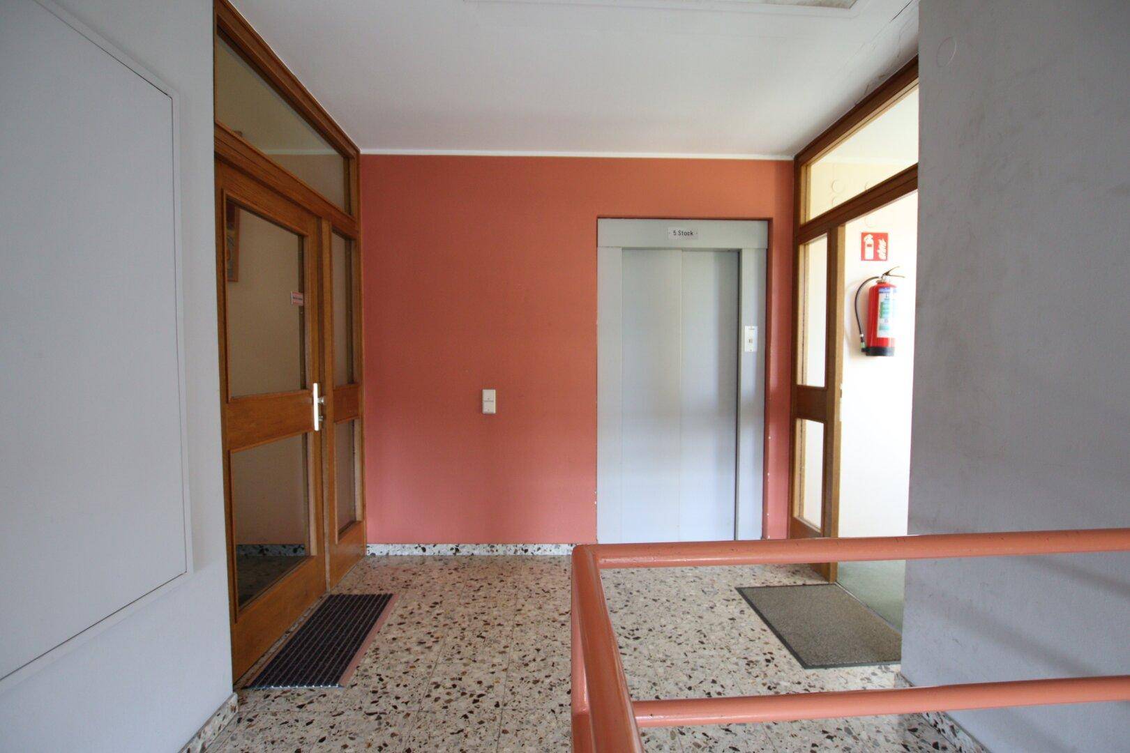 Gemeinschaftlicher Flur mit Aufzug, Terrazzoboden und roter Wandfarbe.