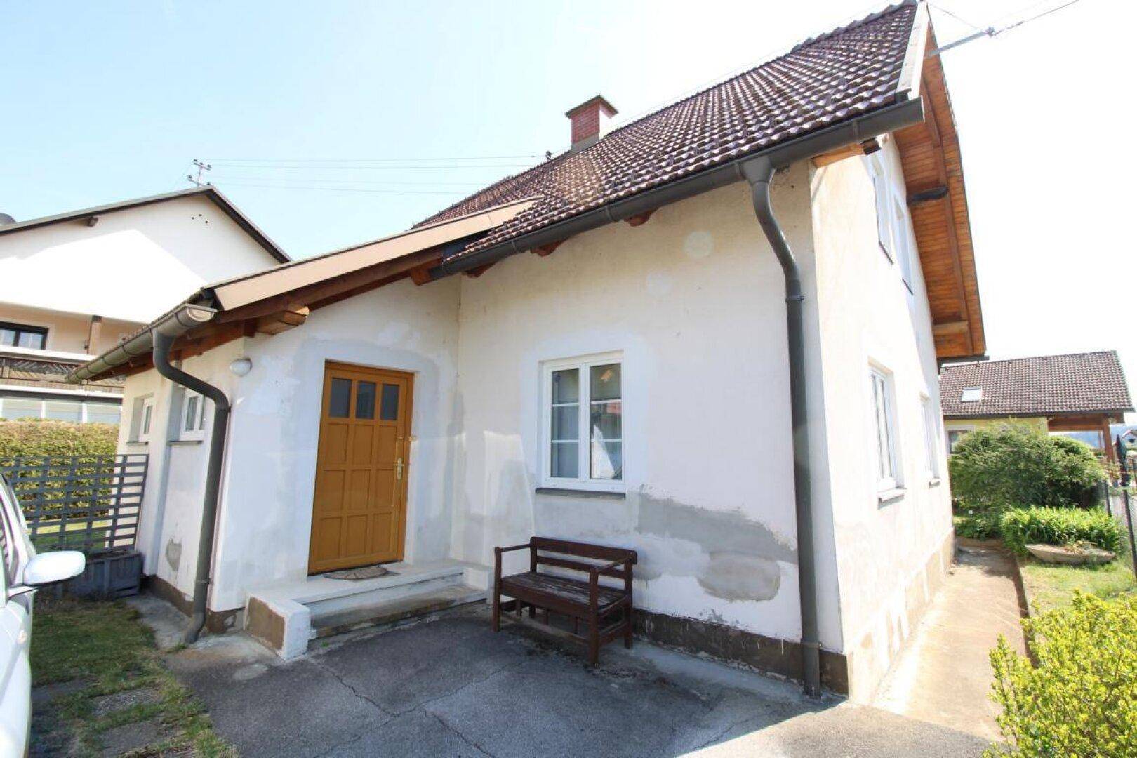 Einfamilienhaus mit heller Fassade, brauner Eingangstür und kleiner Sitzbank im Vorgarten.