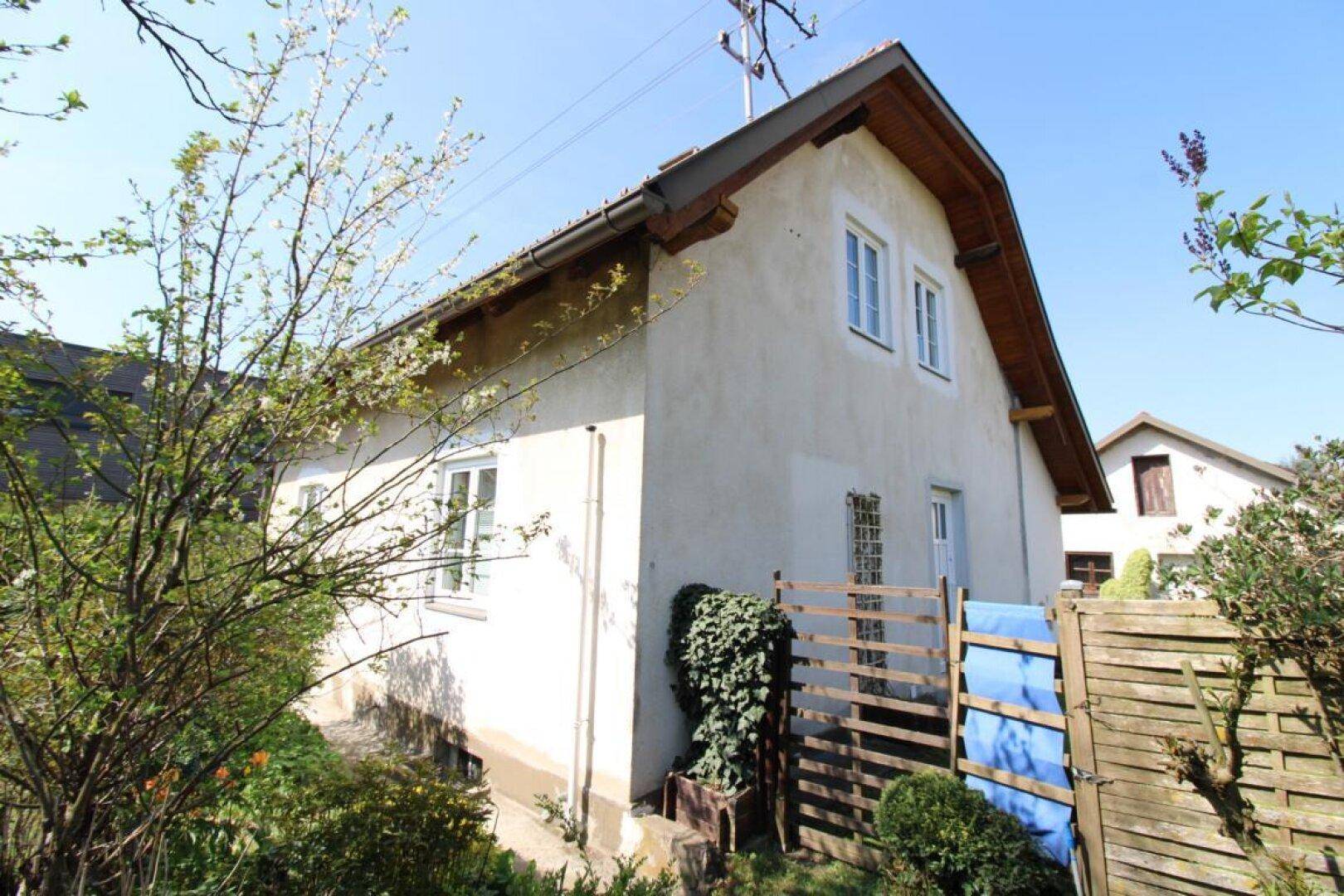 Seitenansicht des Hauses mit Fenstern und einer Holzverkleidung unter dem Dach, umgeben von Bäumen.