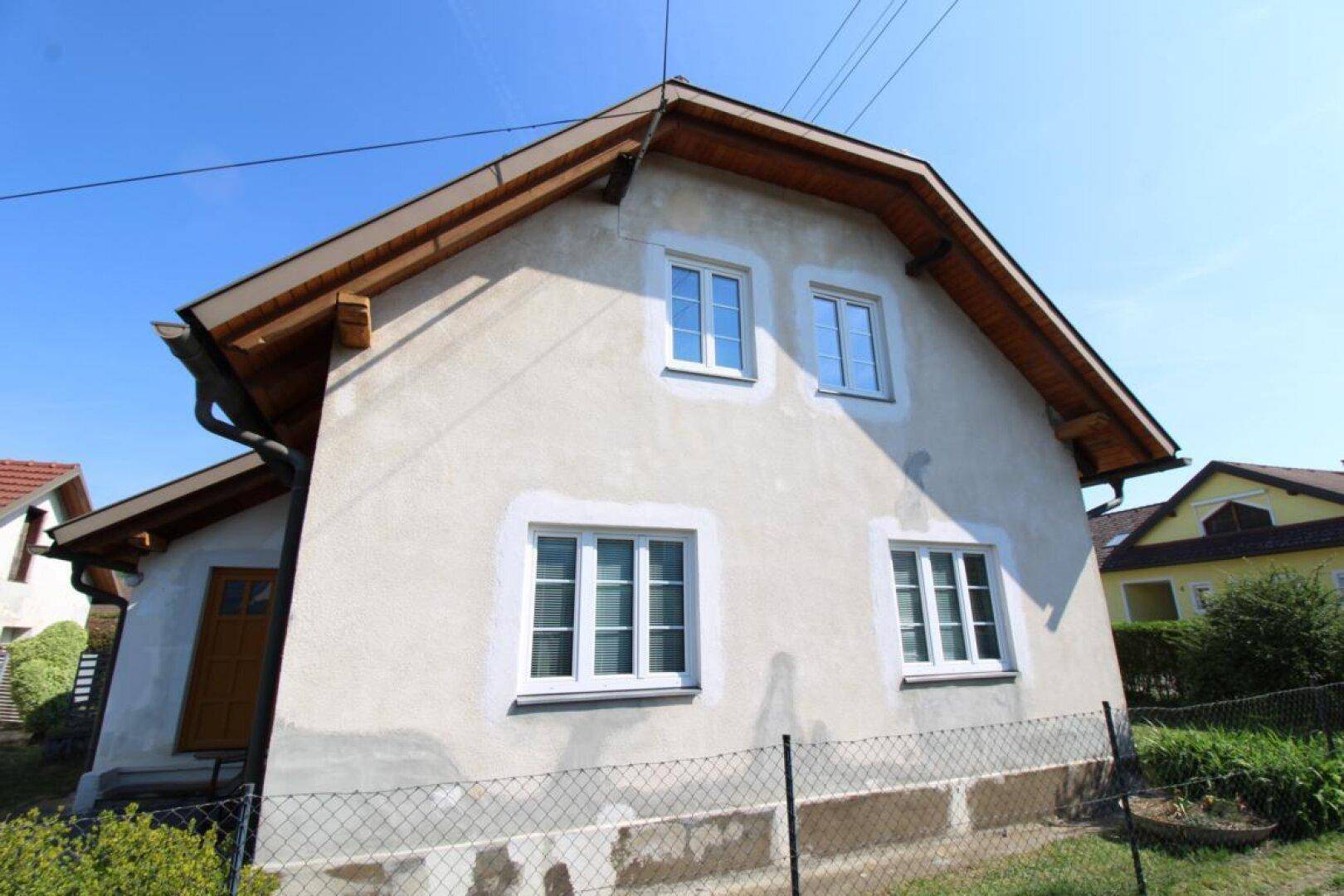 Seitenansicht des Hauses mit weißen Fenstern und einer braunen Holztür, umgeben von Grünflächen.