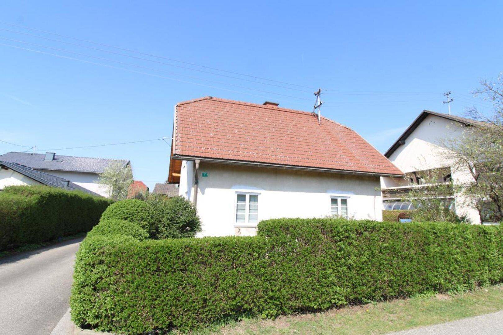 Vorderansicht eines Hauses mit Ziegeldach, umgeben von einer hohen grünen Hecke.