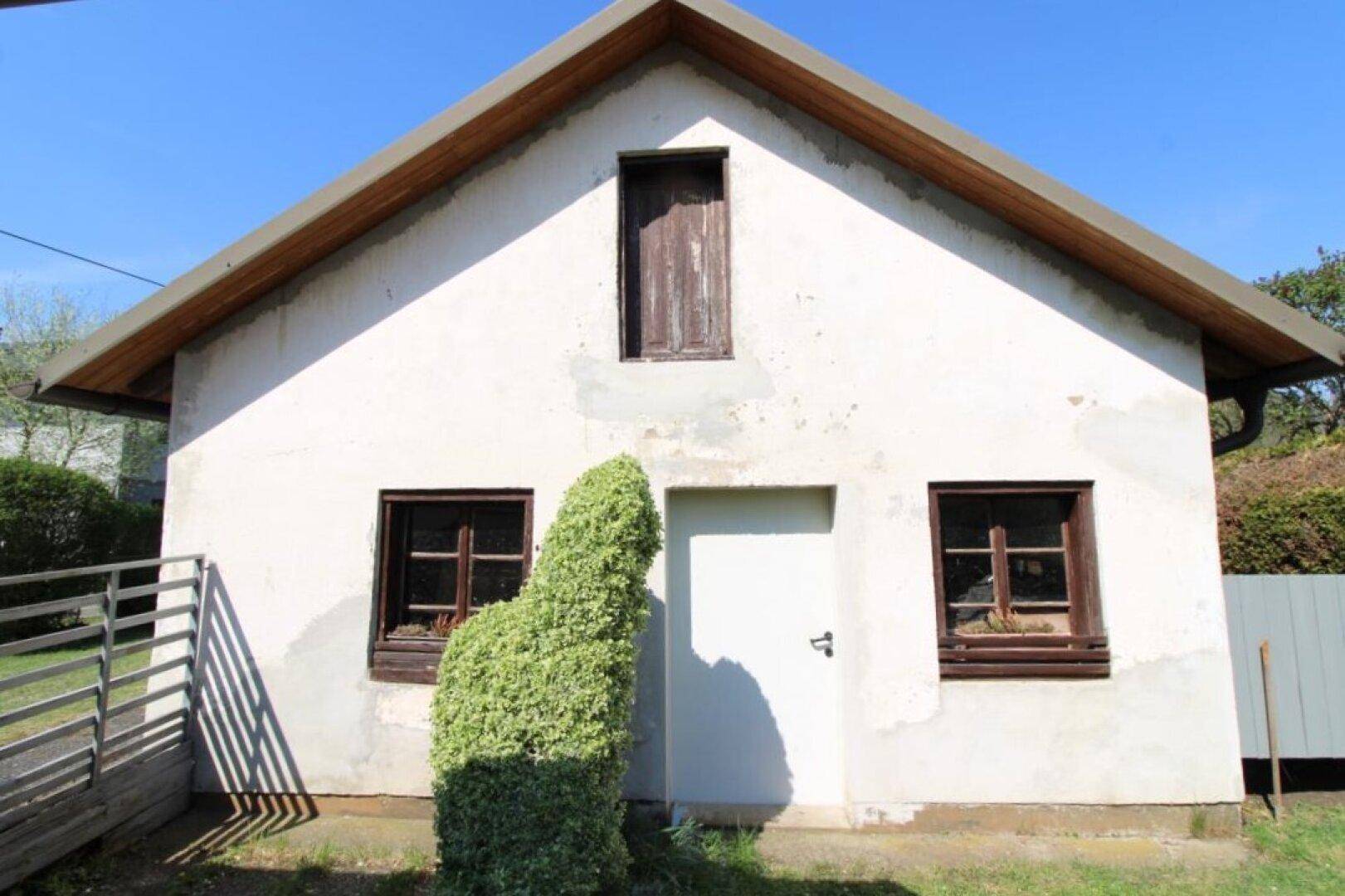 Rückseite des Hauses mit einer weißen Tür und dunklen Fenstern, umgeben von einem Gartenbereich.