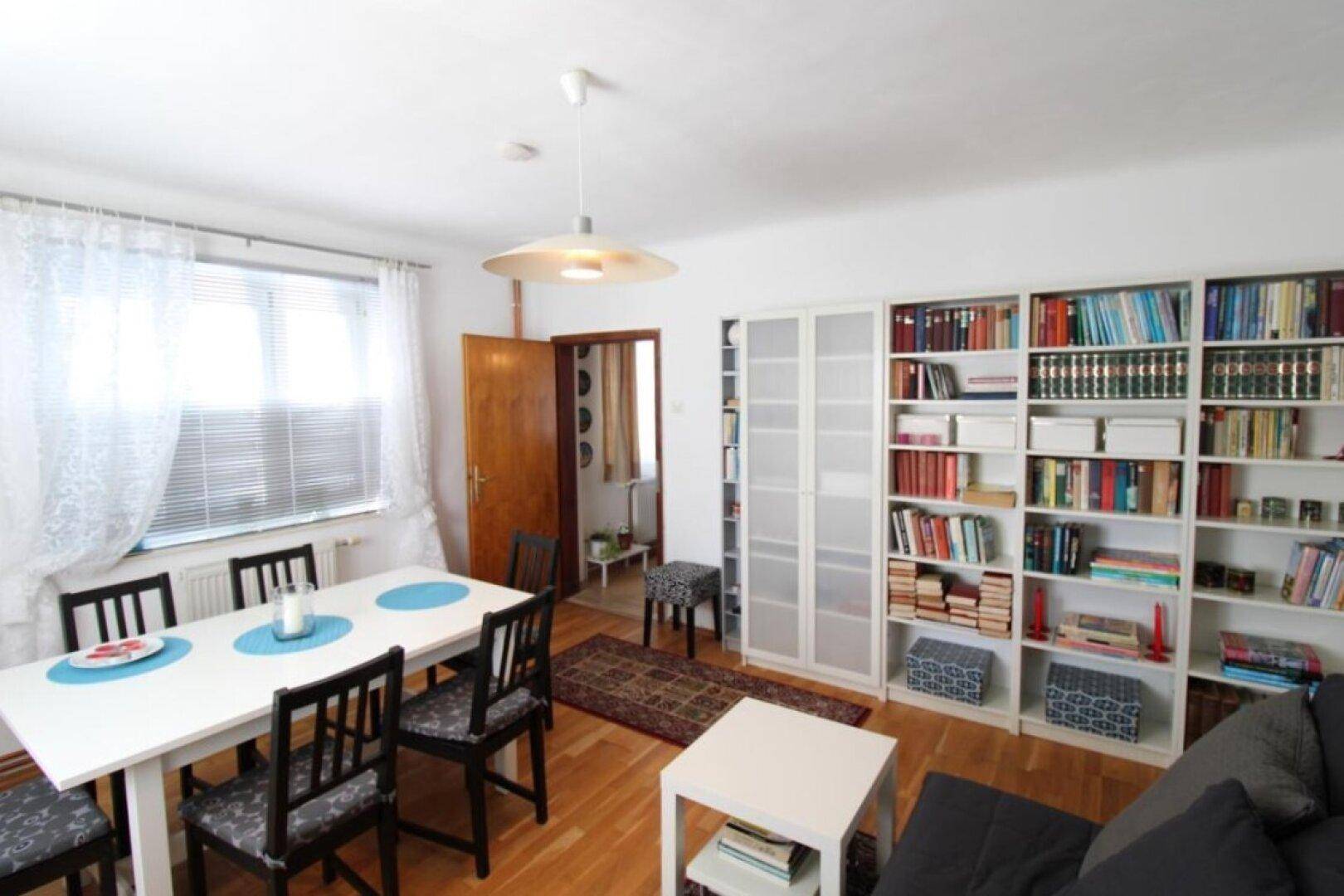 Wohn- und Essbereich mit Bücherregalen, Holzboden und viel Tageslicht durch die Fenster.