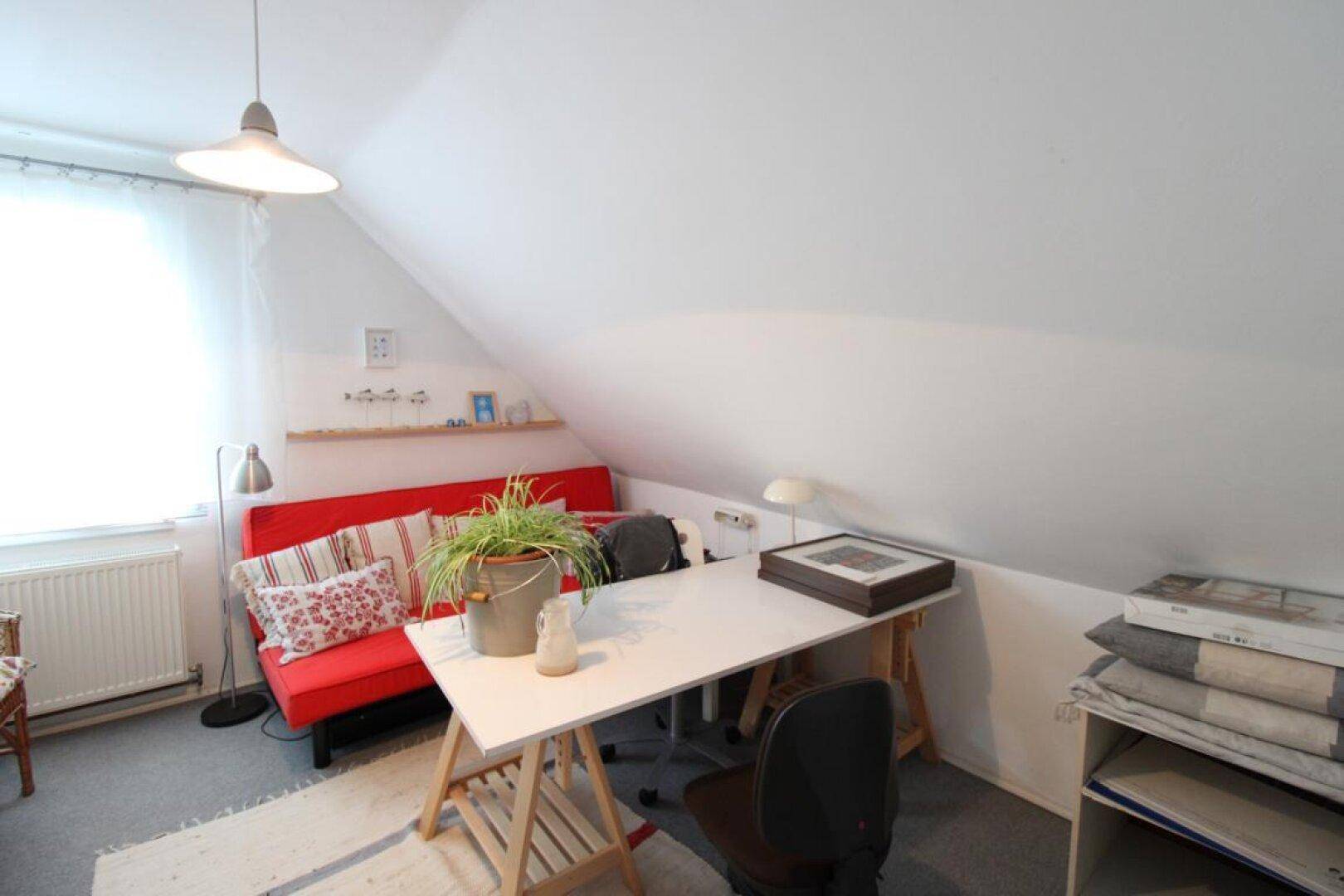 Funktional eingerichtetes Dachzimmer mit Schreibtisch, rotem Sofa und Pflanzen.