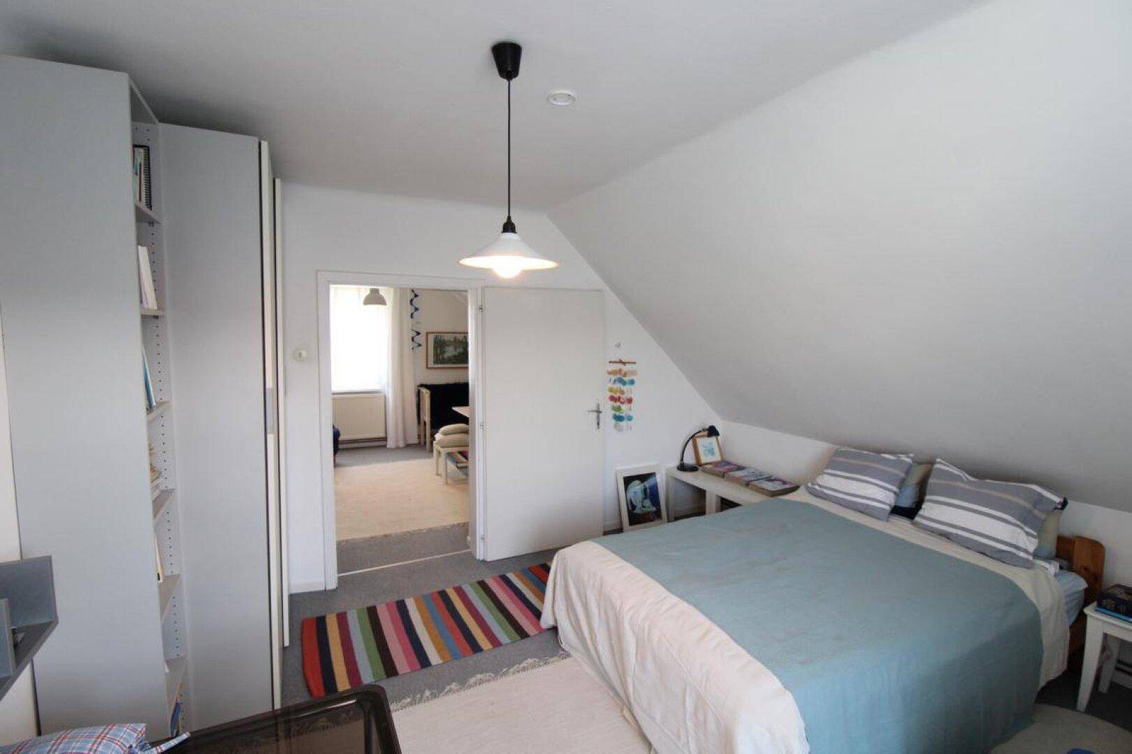 Schlafzimmer im Dachgeschoss mit Bett, Kleiderschrank und Zugang zu einem weiteren Raum.