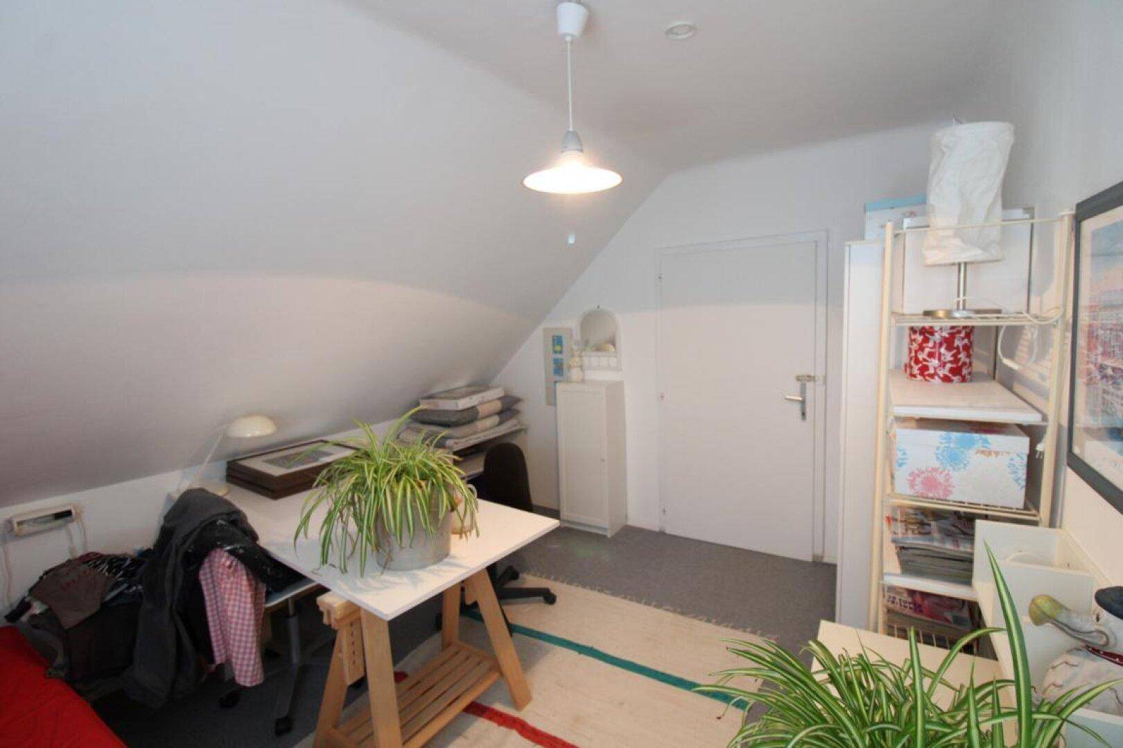 Dachzimmer mit Arbeitsbereich, Stauraum und Zugang zu einem weiteren Raum.