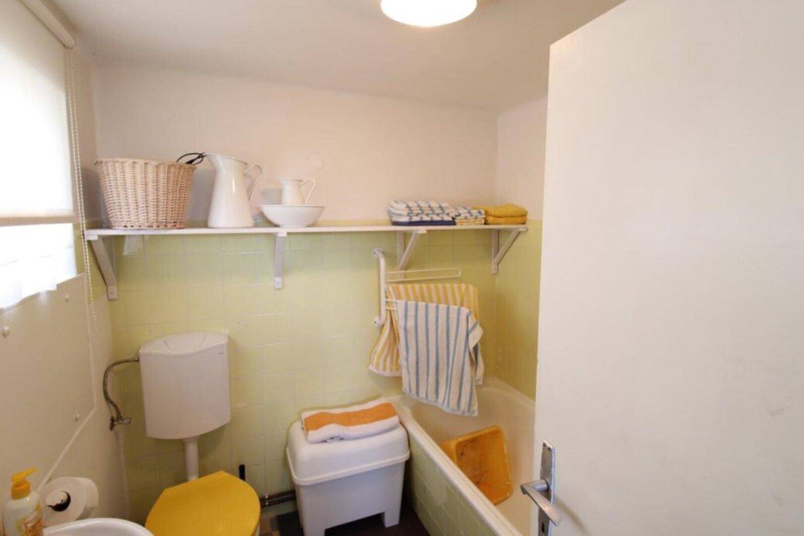 Badezimmer mit Badewanne, Toilette und Fenster, einfache Ausstattung und Ablageflächen.