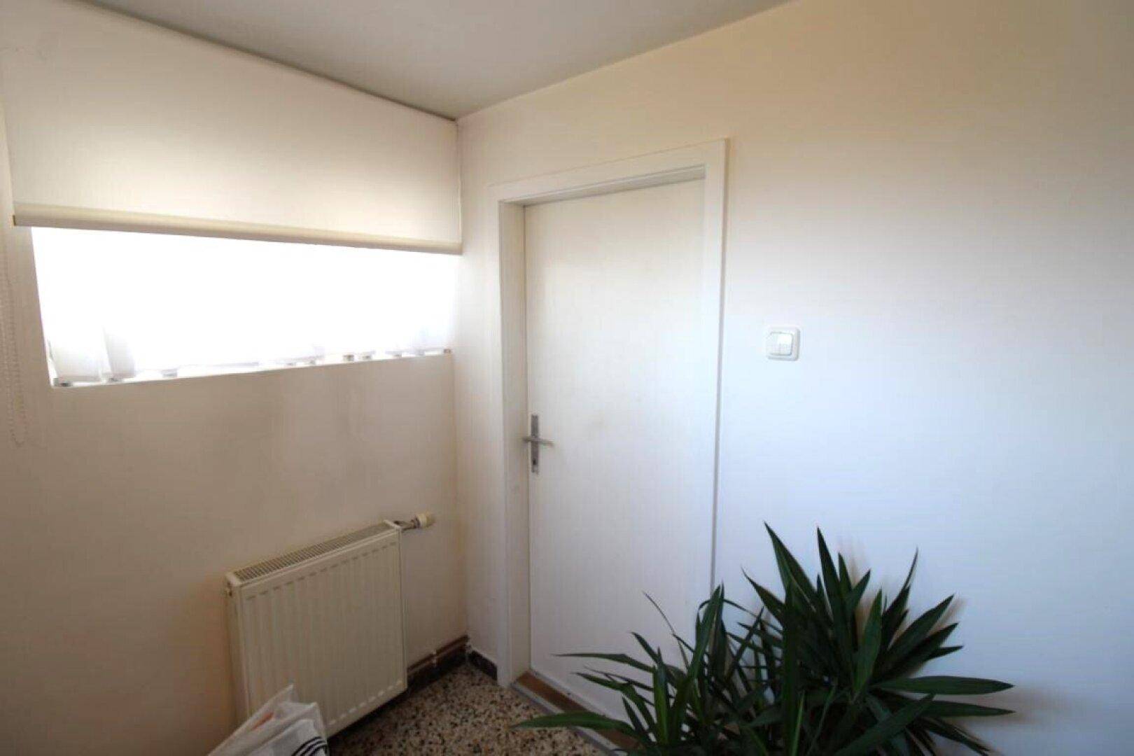 Heller Flur mit Fenster, Heizkörper und einer Pflanze, in einem einfachen Stil gehalten.