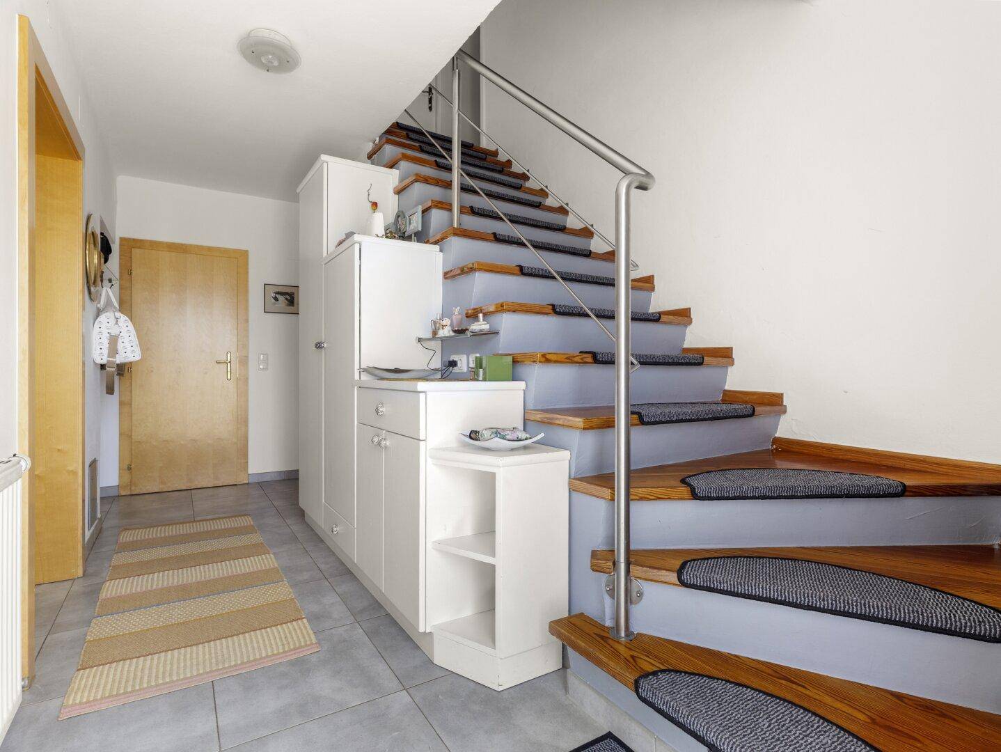 Heller Flur mit Fliesenboden, Holztreppe und integriertem Stauraum unter der Treppe.