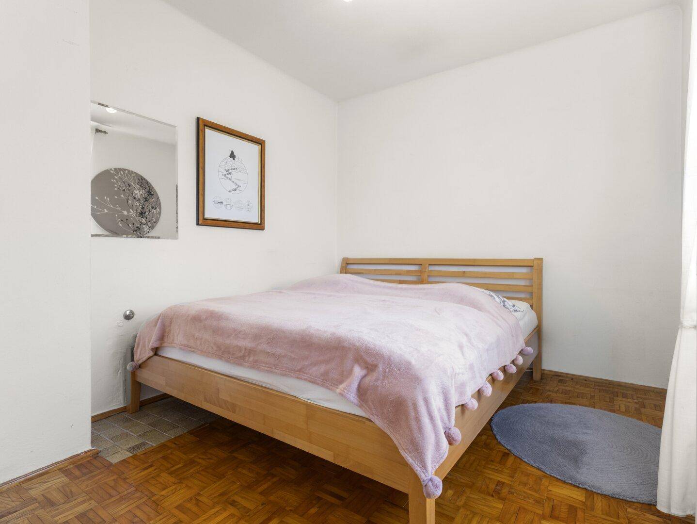Einfaches Schlafzimmer mit Holzbett, Parkettboden und einem kleinen Fenster.
