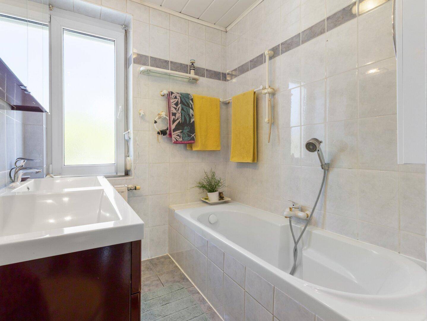 Gefliestes Badezimmer mit Badewanne, Einzelwaschbecken und Fenster für natürliche Belüftung.