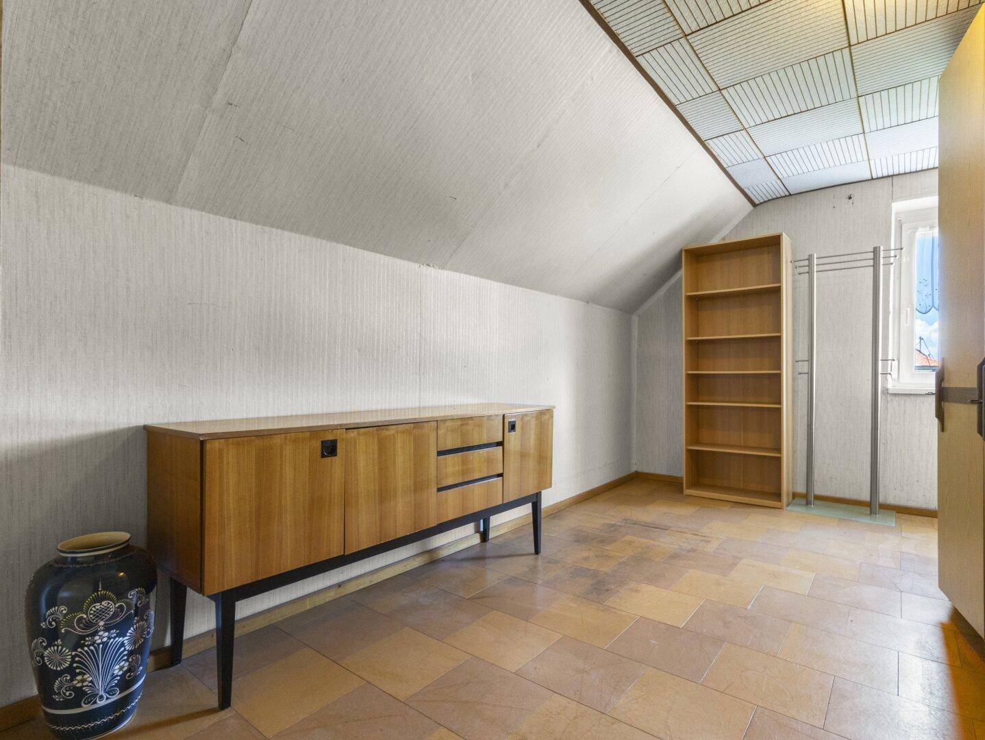Helles Dachzimmer mit schräger Decke, Holzkommode und Fliesenboden, ideal als zusätzlicher Raum.