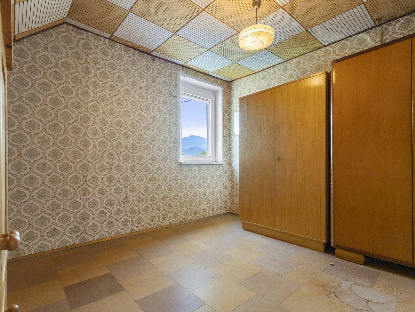 Schlafzimmer mit gemusterter Tapete, Holzschränken und einem Fenster mit Bergblick.