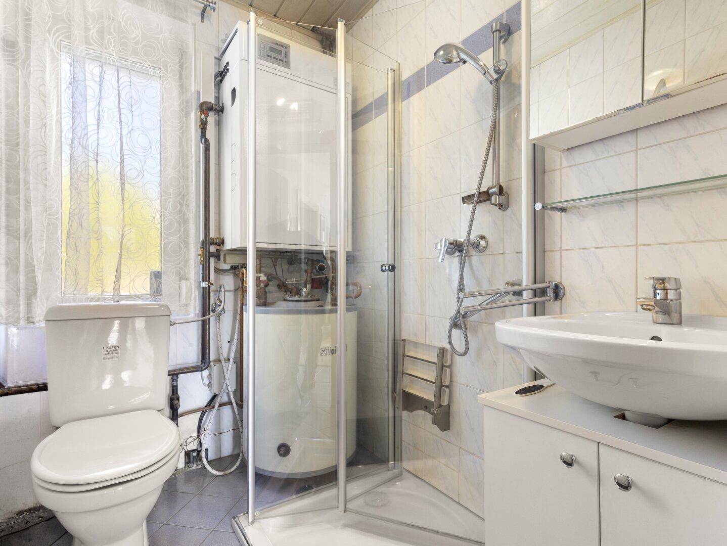 Funktionales Badezimmer mit Toilette, Waschbecken, Duschkabine und einem sichtbaren Warmwasserbereiter.