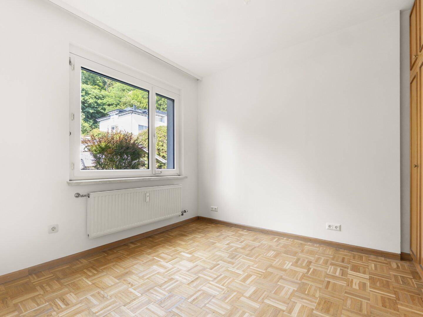 Helles Schlafzimmer mit Parkettboden, weißen Wänden und einem Fenster mit Blick ins Grüne.