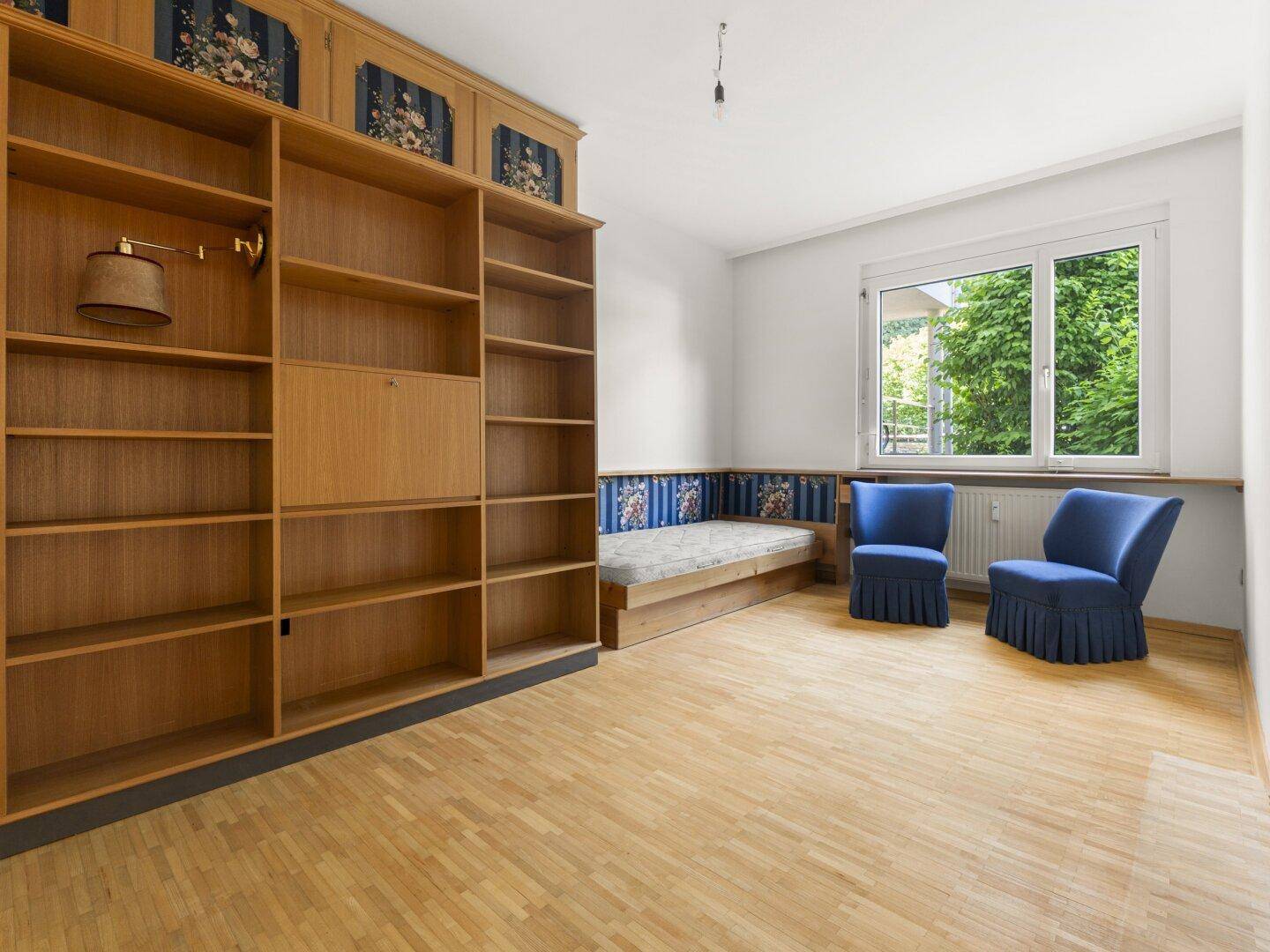 Geräumiges Schlafzimmer mit großem Holzregal, Tagesbett und zwei blauen Sesseln, Parkettboden.