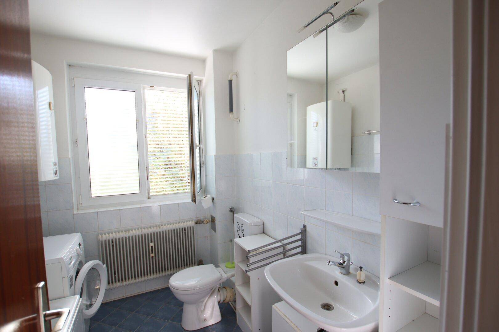 Helles Badezimmer mit Fenster, WC, Waschbecken und Waschmaschine, funktional und gut beleuchtet.