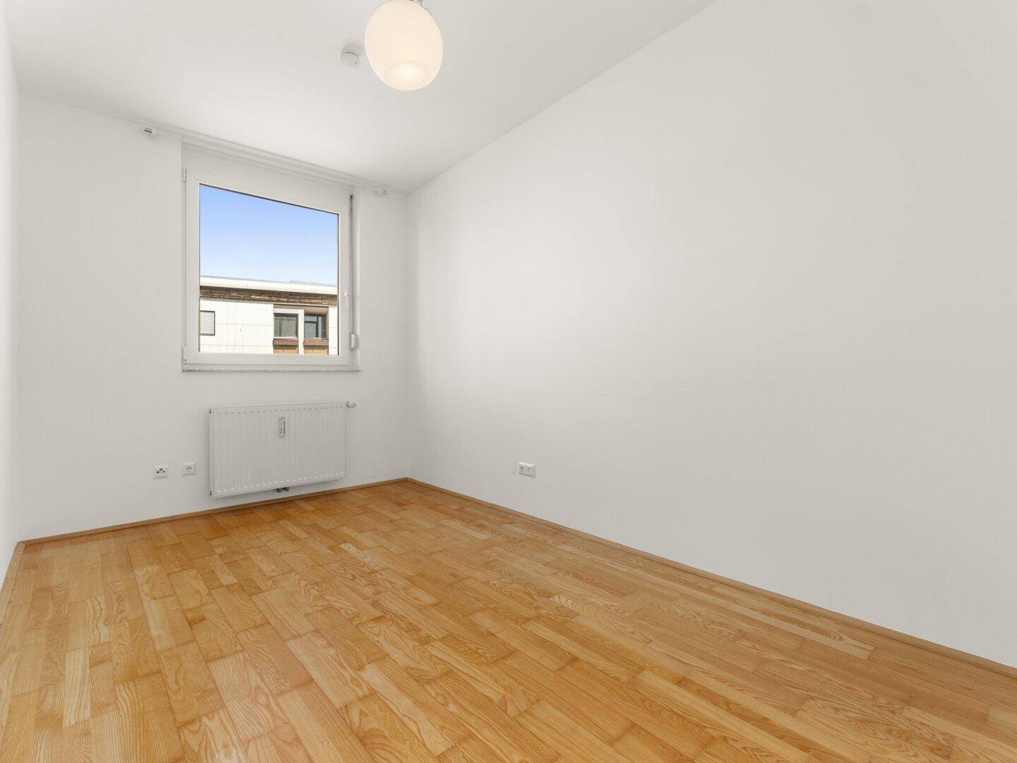 Helles Schlafzimmer mit Parkettboden und einem Fenster, das viel Licht hereinlässt.