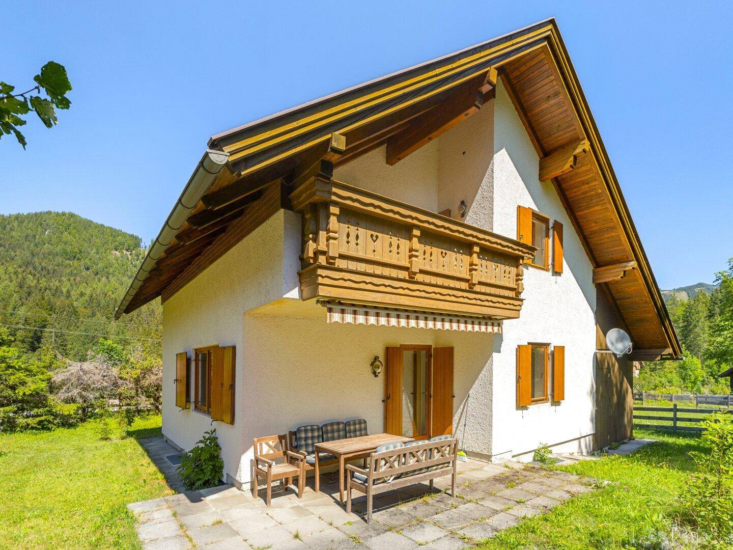 Hausfassade mit kleiner Terrasse und traditionellem Holzbalkon, umgeben von grüner Natur.
