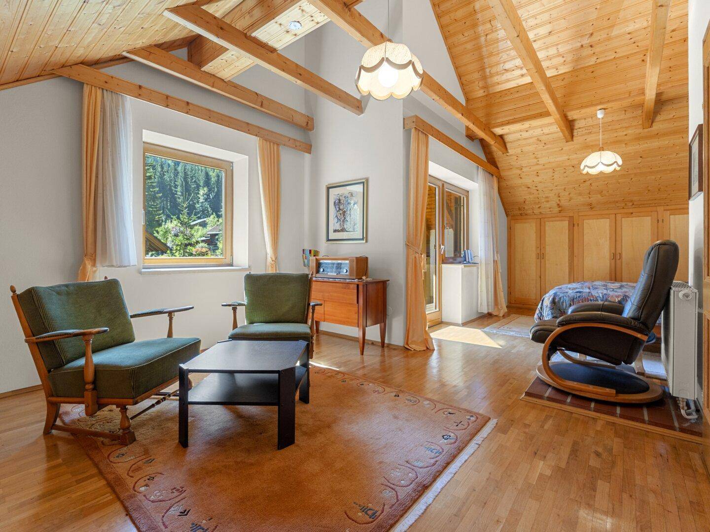 Geräumiges Schlafzimmer mit Holzbalkendecke, Sitzbereich und Blick ins Grüne.