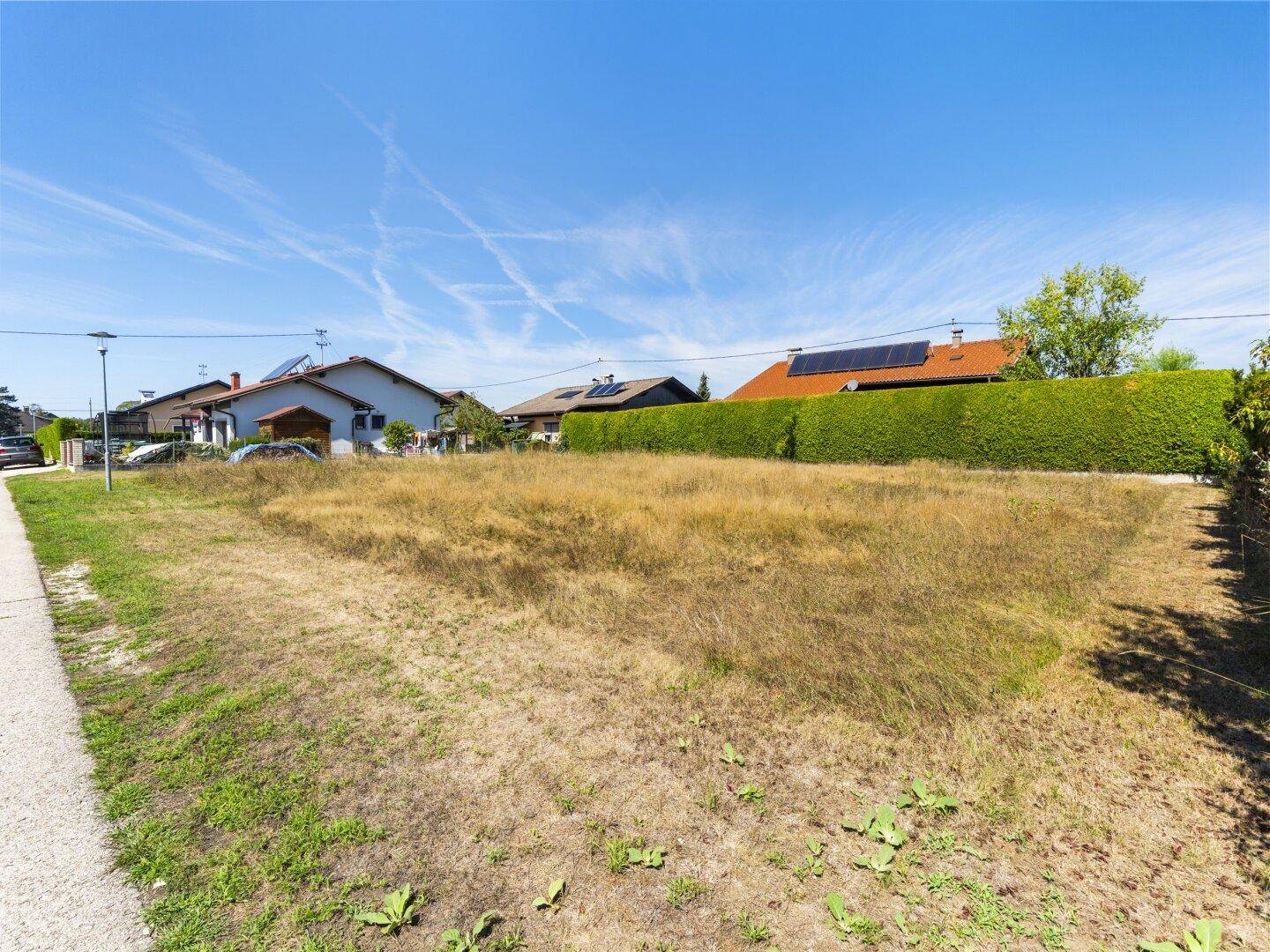 Grundstück mit trockenem Gras, angrenzendem Gehweg und Wohnhäusern im Hintergrund unter blauem Himmel.