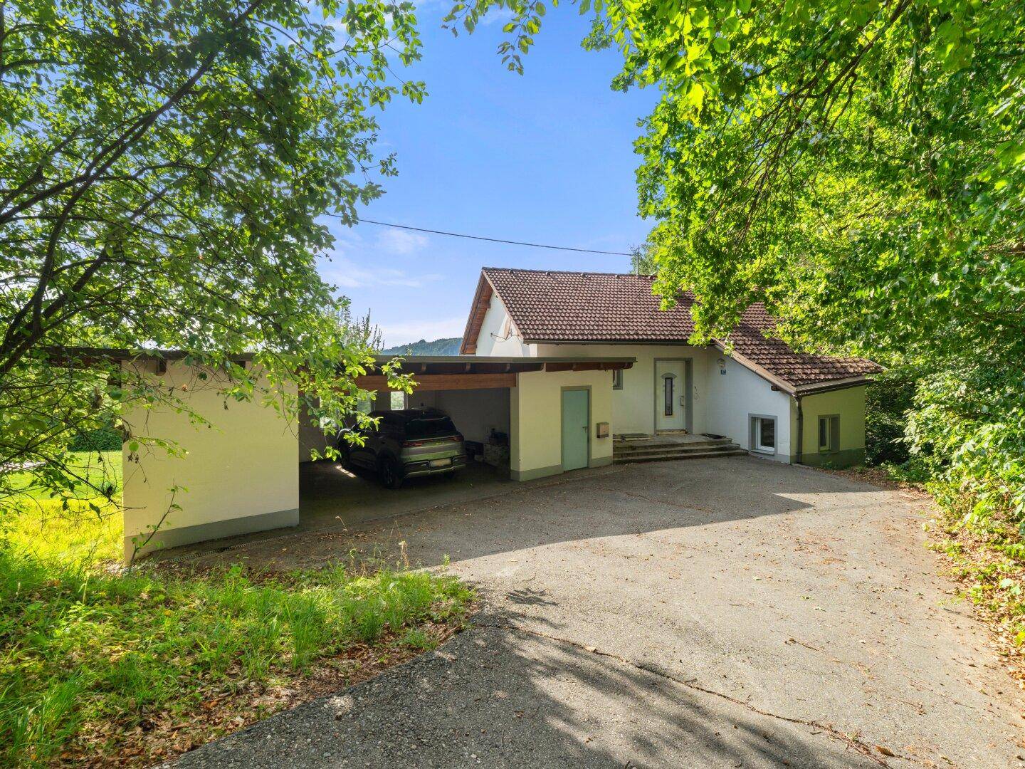 Vorderansicht des Hauses mit Carport und Zufahrt, eingebettet in eine grüne, bewaldete Umgebung.