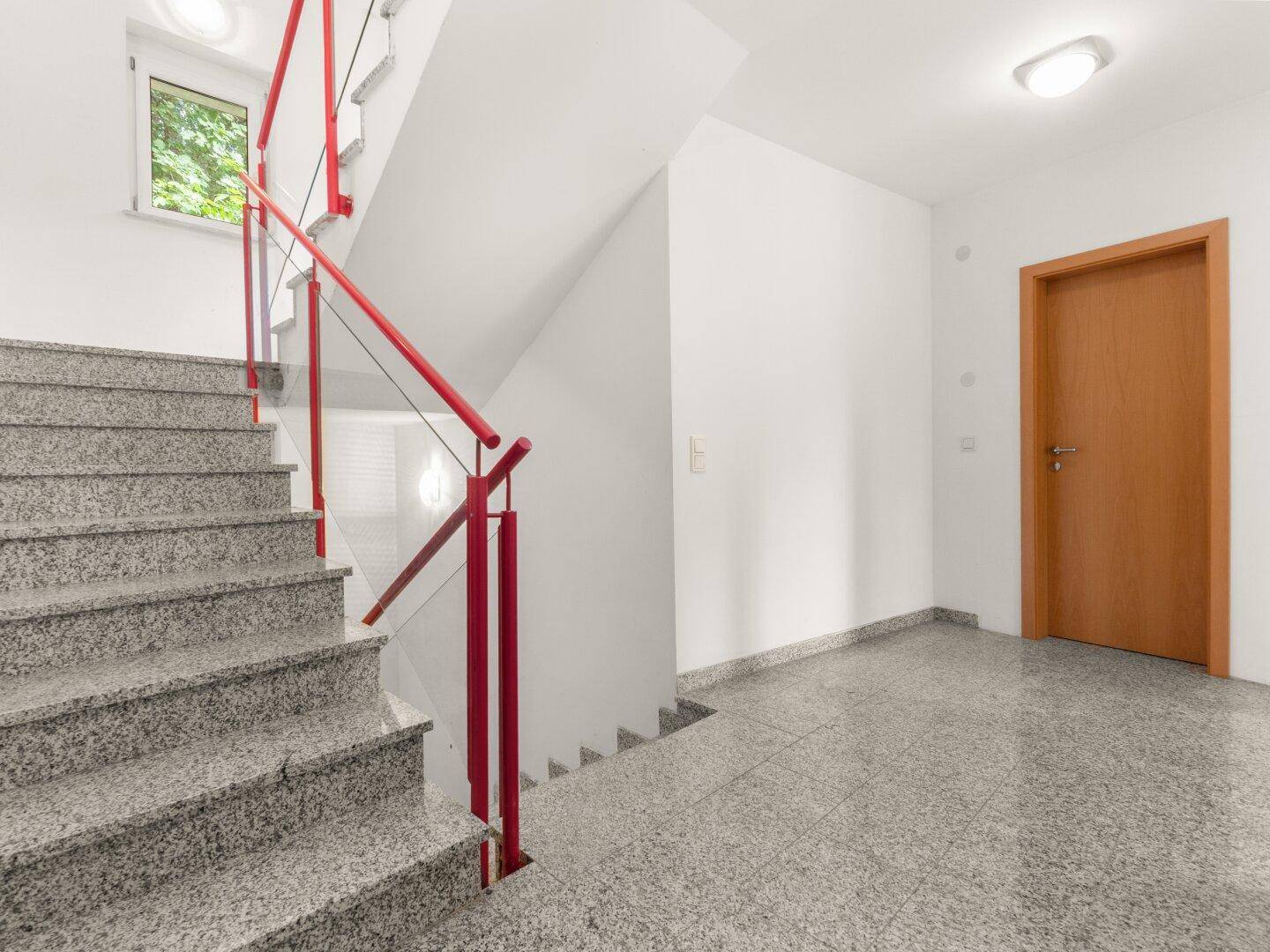 Heller Treppenaufgang mit Granitboden und rotem Geländer, führt zu einer Holztür im Flur.