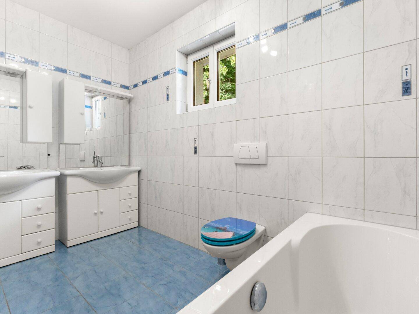 Gefliestes Badezimmer mit Badewanne, Doppelwaschbecken und Fenster, in hellen Farben gehalten.