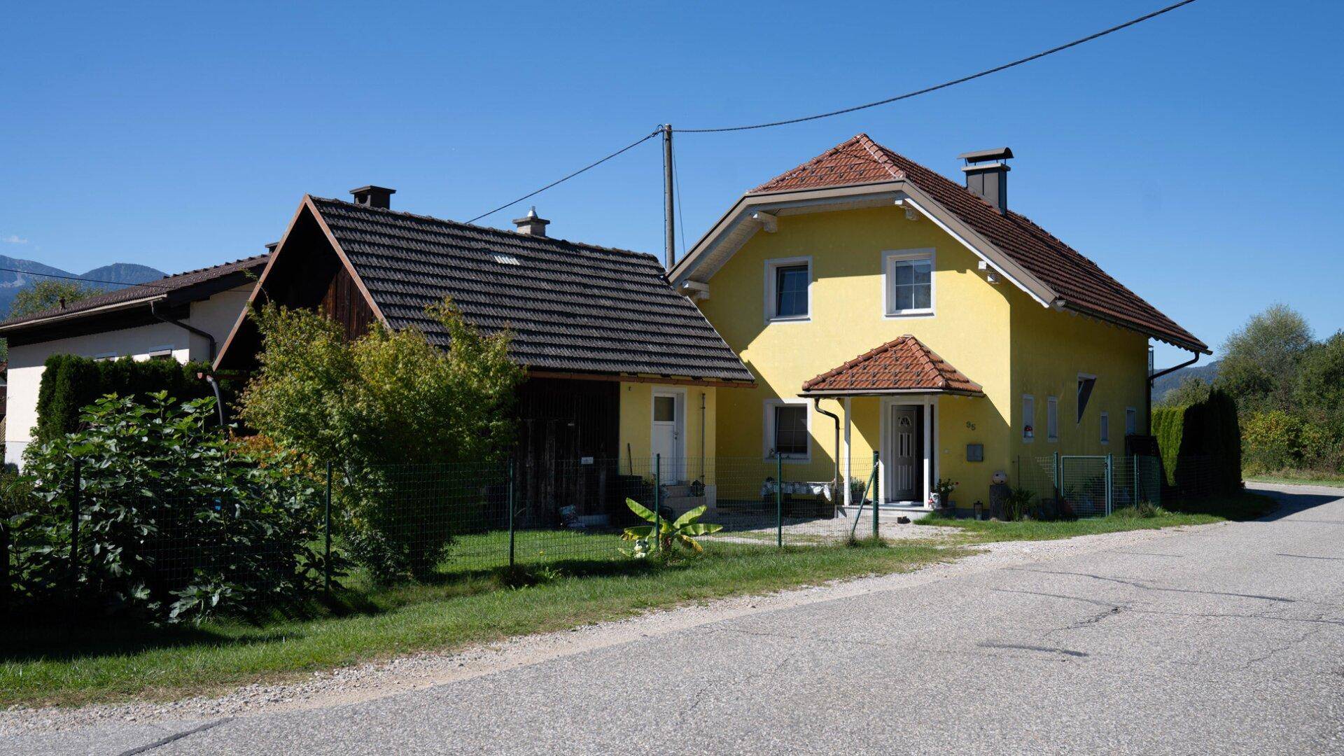 Ansicht des gelben Hauses mit rotem Ziegeldach und einem angrenzenden Holzschuppen unter blauem Himmel.