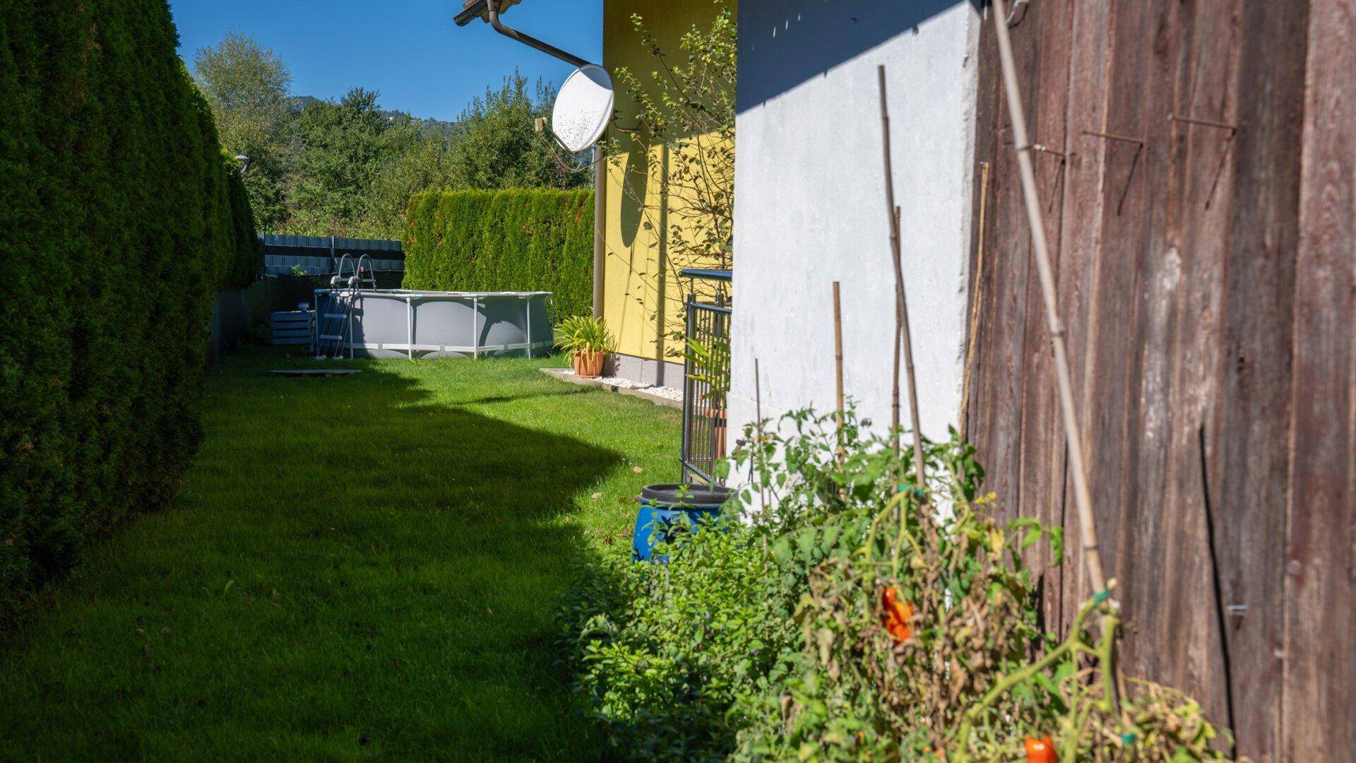 Seitlicher Gartenbereich mit Rasen, Hecke und einem Aufstellpool im Hintergrund.